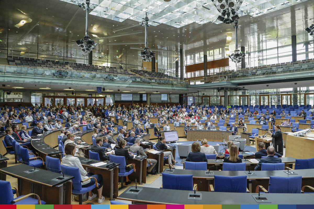 Auf dem Bild sind die kreisförmig ausgerichteten Stuhlreihen eines Plenums zu sehen. Einige der Stühle sind frei, der Rest ist von Menschen besetzt. In der Mitte des Plenums steht eine Frau am Rednerpult und spricht zu den versammelten Menschen.