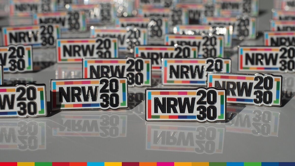 Mehrere Anstecker mit dem Logo von NRW2030 auf einer verspiegelten Fläche