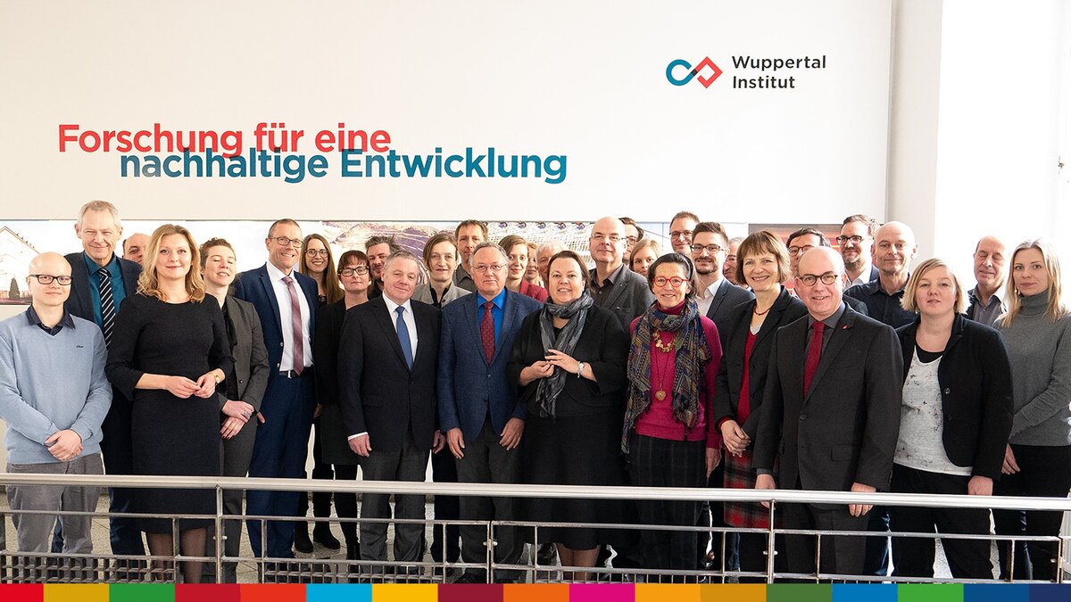 Das Bild zeigt die Mitglieder des Teams Nachhaltigkeit. Sie stehen in mehreren Reihen und blicken in die Kamera. Im Hintergrund eine Wand. Auf dieser Wand ist das Logo der Universität Wuppertal zu sehen und darunter ist der Slogan "Forschung für einen nachhaltige Entwicklung" zu lesen. 